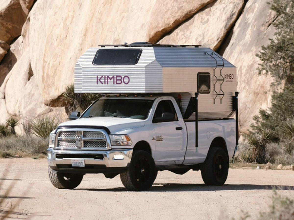 White Dodge Ram 2500 with Kimbo 8 hard-side aluminum truck bed camper at a Joshua Tree National Park granite-boulder pull-off, photographed front-quarter so the truck face and the camper KIMBO wordmark are both visible.
