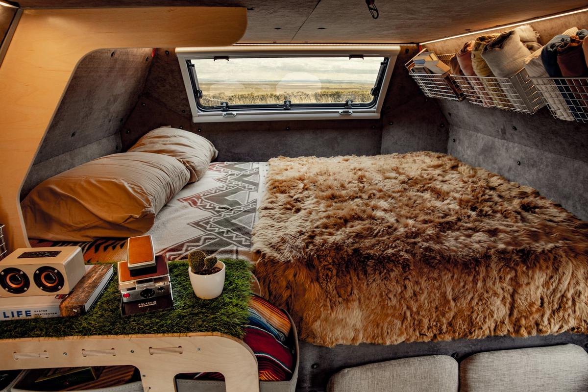 Cozy cabover sleeping nook of a Kimbo aluminum truck bed camper with bamboo trim, sheepskin throw, cushioned pillows, a vintage Polaroid camera and a small cactus on the side shelf, the picture window overlooking distant mountains.