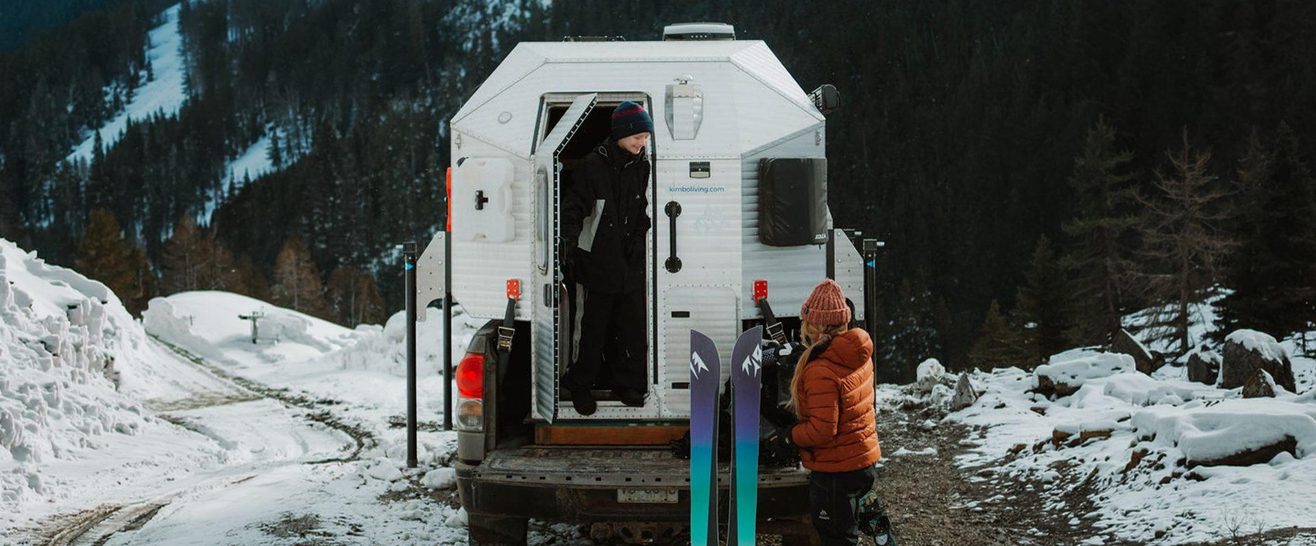 Kimbo owner standing in the open rear doorway of a white Kimbo hard-side truck bed camper at a snowy Pacific Northwest pull-off, skis racked along the side and a friend in a knit cap waiting outside in the snow.