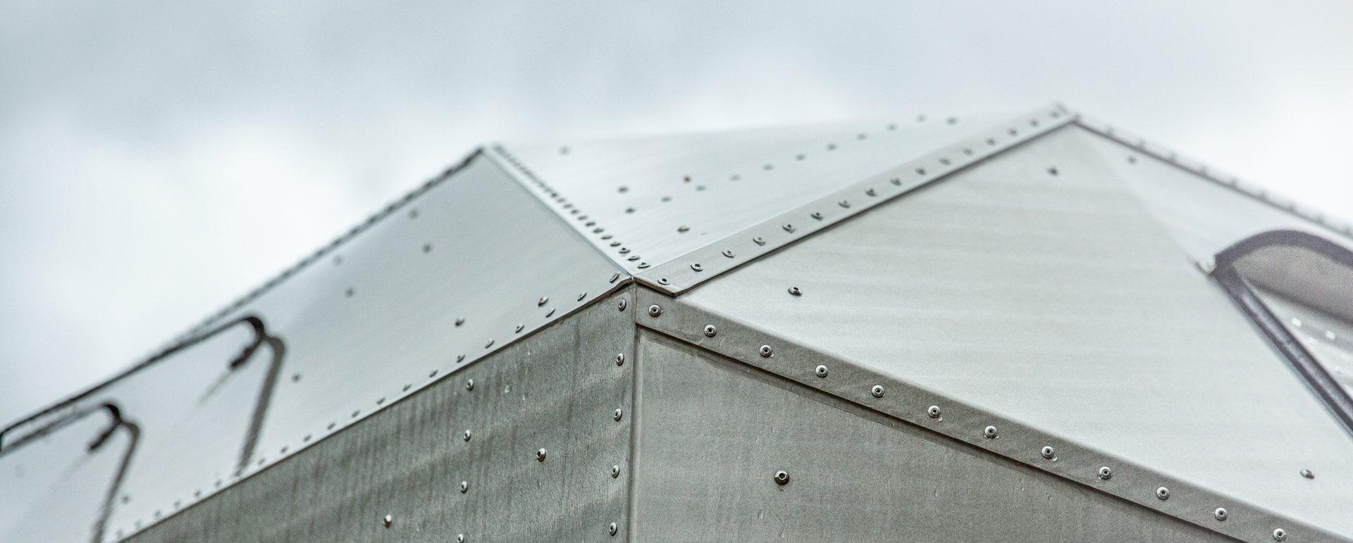 Close-up of the hand-riveted aluminum roof apex of a Kimbo hard-side truck bed camper, showing the laser-cut 5052 panels seamed together with solid rivets at 1.5″ spacing and the integrated black side ladder.