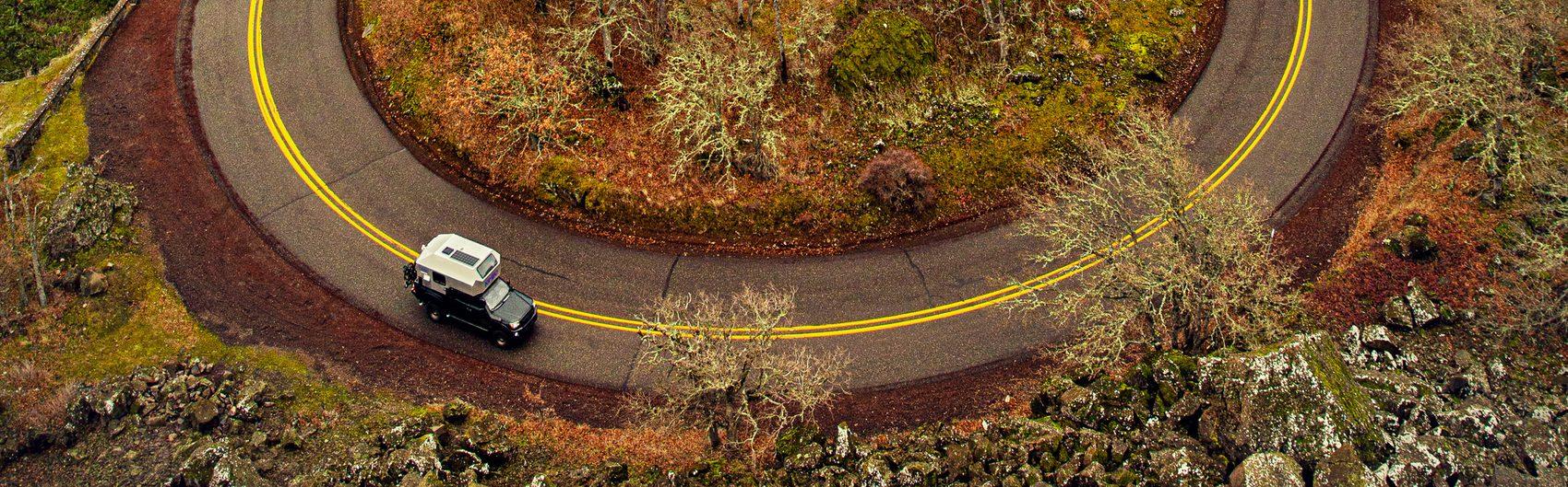 Aerial drone view of a Pacific Northwest highway switchback ringed with yellow centre-line striping, a white pickup with Kimbo hard-side slide-in truck bed camper driving the loop, autumn vegetation and basalt rock outcroppings filling the surrounding terrain.