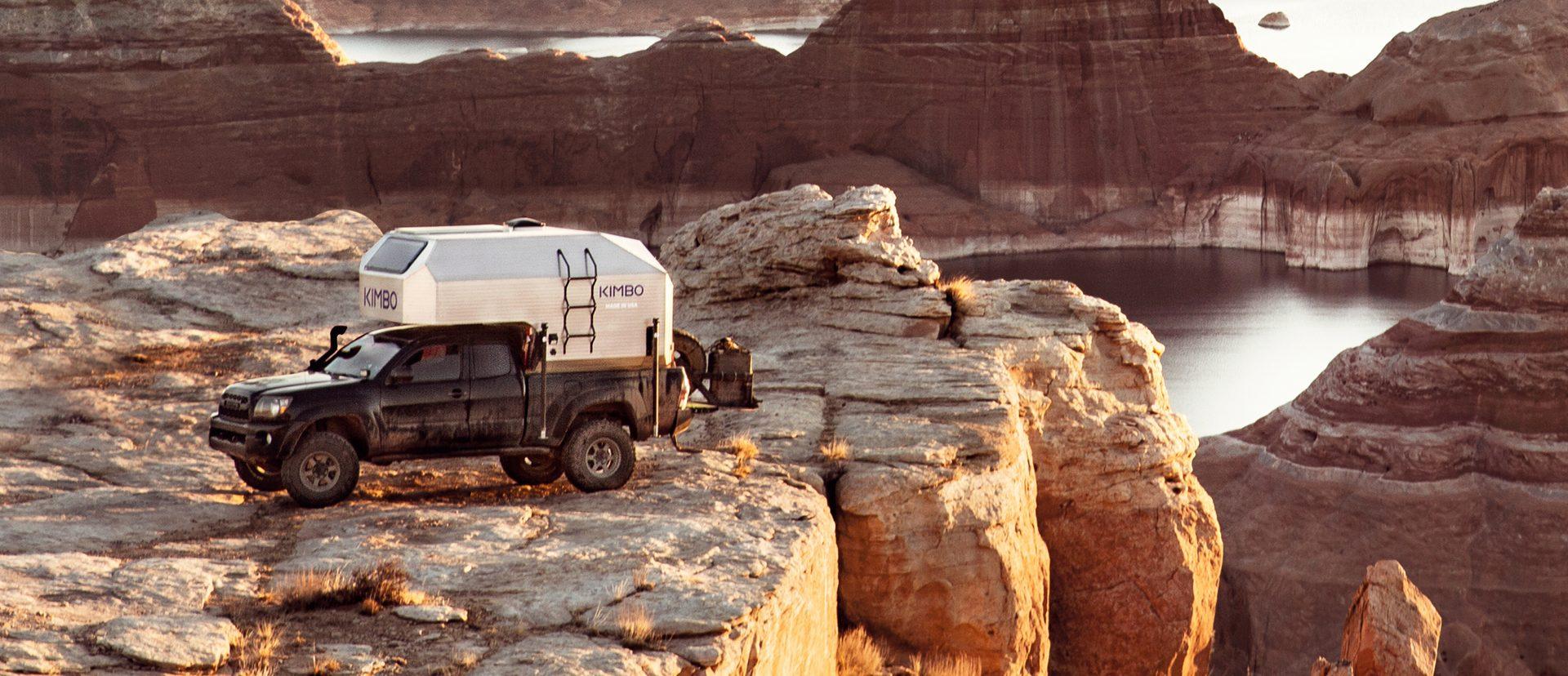Black Toyota Tacoma with Kimbo 6 hard-side slide-in truck bed camper parked on red sandstone above a Lake Powell side canyon at golden hour, southern Utah.