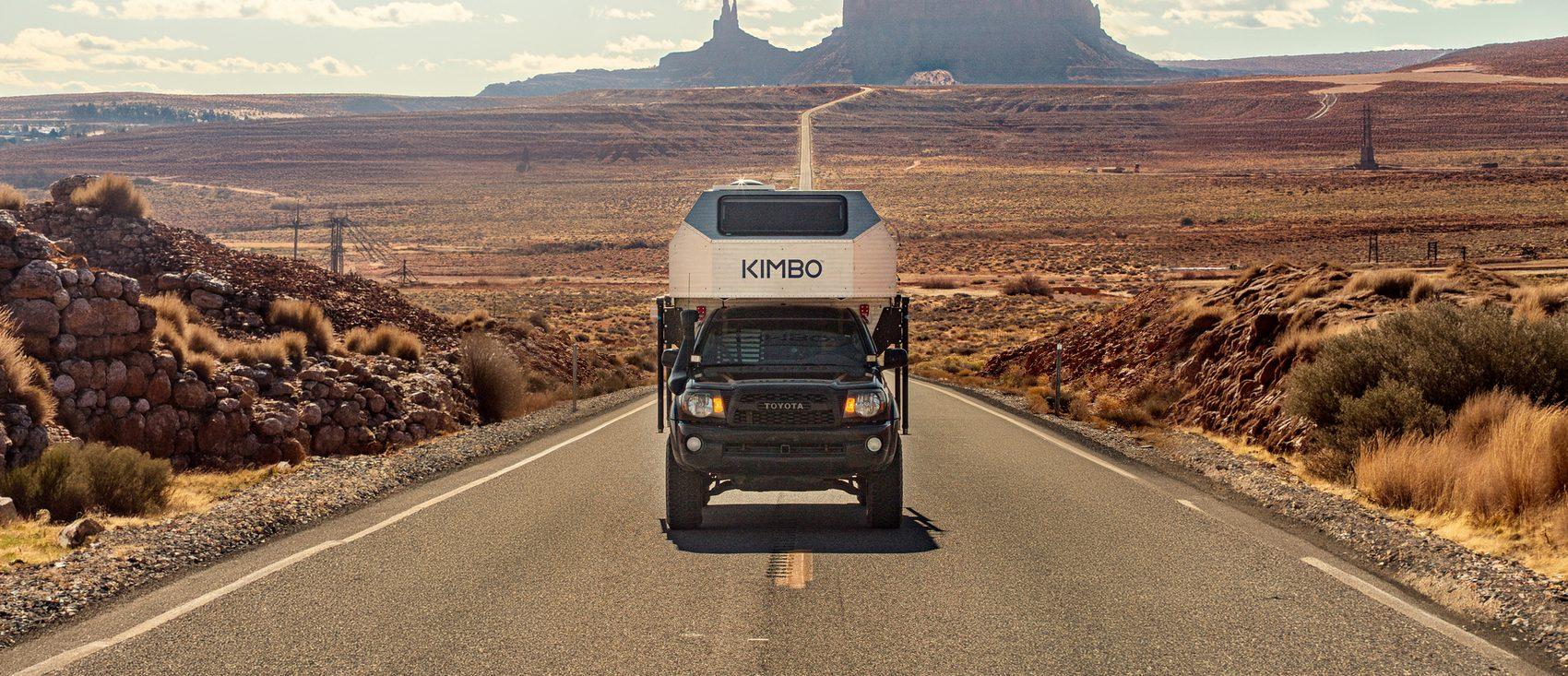 Black Toyota Tacoma with Kimbo 6 hard-side slide-in truck bed camper driving an empty desert highway toward the Monument Valley sandstone monoliths at sunset, southwest Utah.