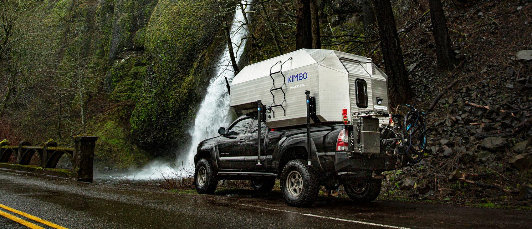 Black Toyota Tacoma with Kimbo 6 hard-side slide-in truck bed camper parked beside a roaring Columbia River Gorge waterfall, a mountain bike strapped to the rear rack on a wet Pacific Northwest road.