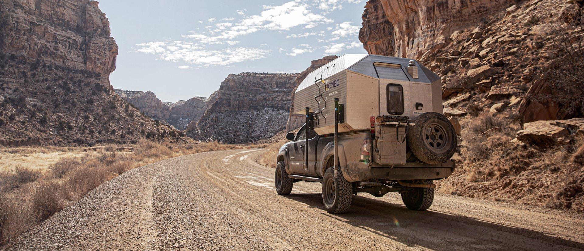 Toyota Tacoma with Kimbo 6 hand-riveted slide-in truck bed camper driving down a red-rock canyon dirt road in the Utah desert — sandstone walls on both sides, open blue sky and high clouds overhead.