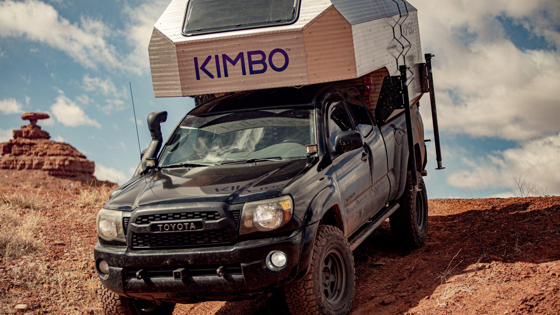 Black Toyota Tacoma with the Kimbo 6 hard-side aluminum truck bed camper installed, photographed front-quarter from a low angle on red sandstone in southern Utah, the Mexican Hat formation visible on the horizon and the KIMBO wordmark prominent on the cabover.