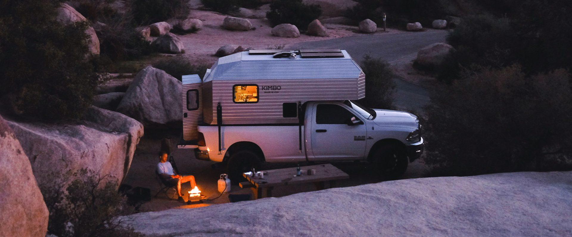 White Dodge Ram 2500 with Kimbo 8 hard-side aluminum truck bed camper boondocking among Joshua Tree granite boulders at dusk, owner seated next to a small campfire and a wooden picnic table, warm interior lights glowing through the windows.